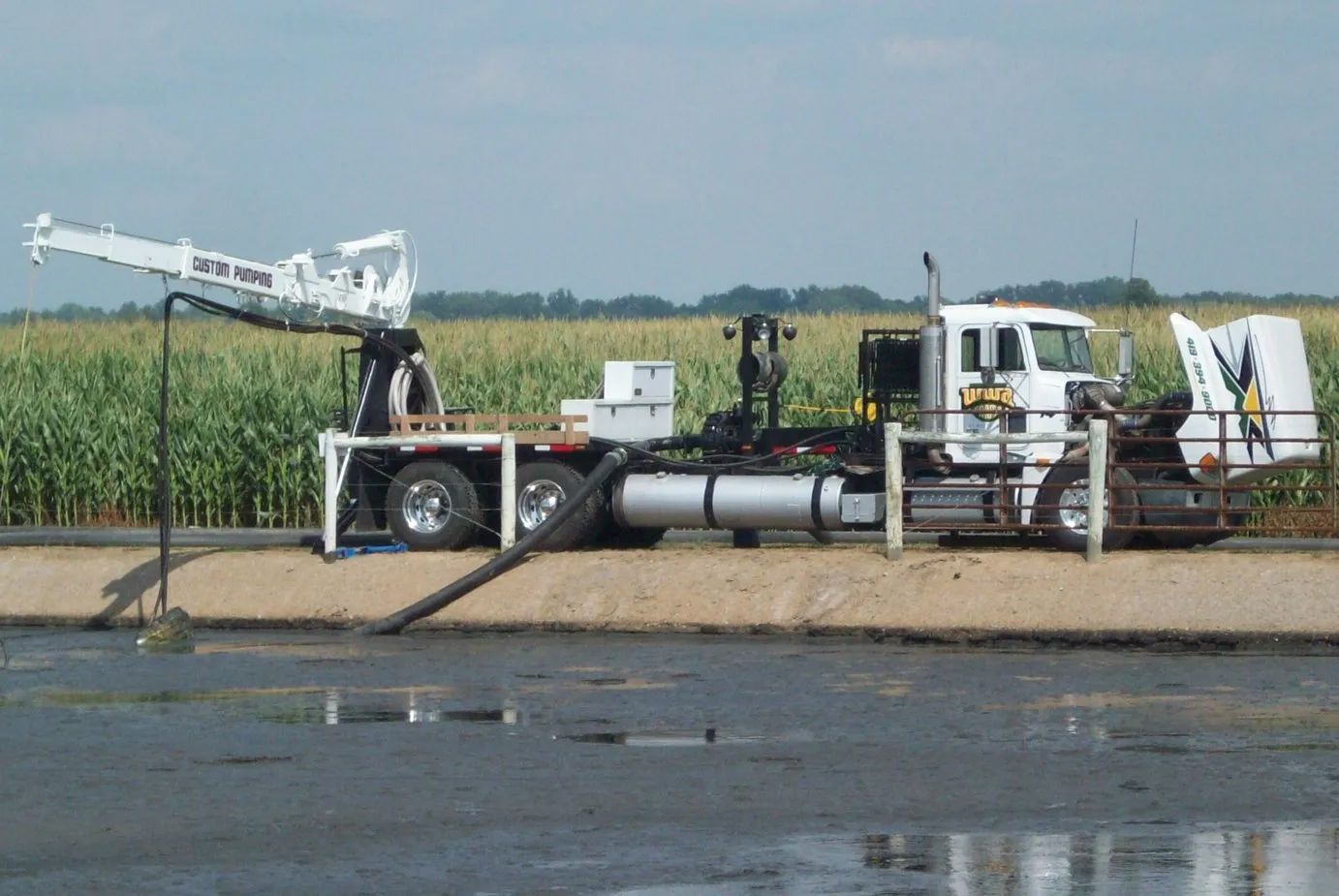 Industrial water-treatment truck beside a canal, with pipes and equipment on a dirt embankment near crops