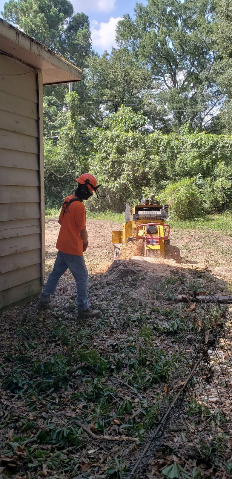 A worker in an orange shirt and hard hat operates a wood chipper on a sunny day next to a house exterior.