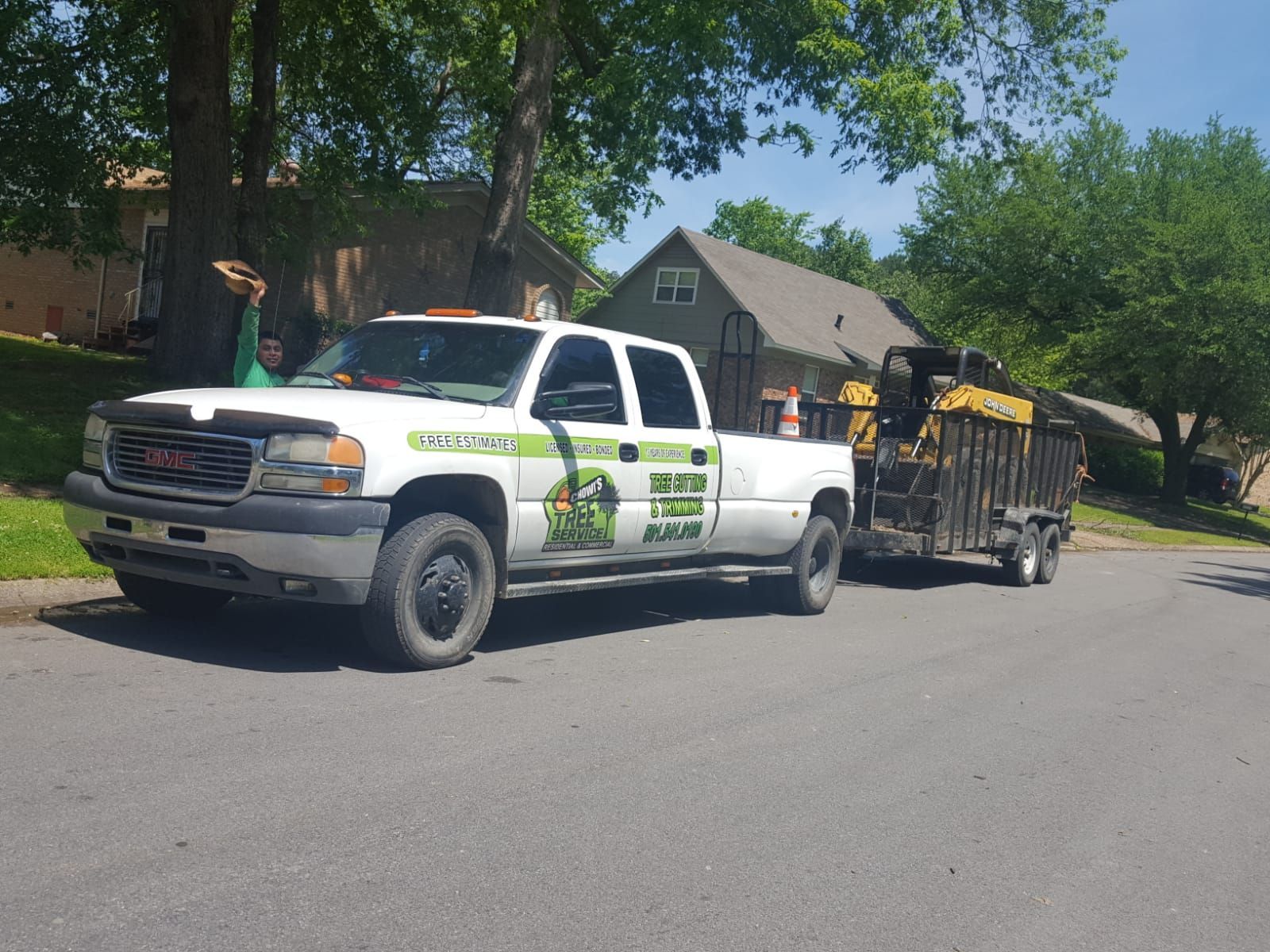 A white GMC work truck towing a trailer with a yellow piece of heavy machinery, parked on a suburban street.