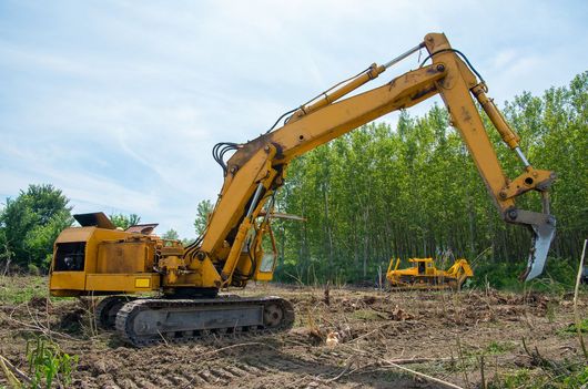 Yellow excavator on caterpillar tracks in a field, with a second piece of construction equipment in the background.