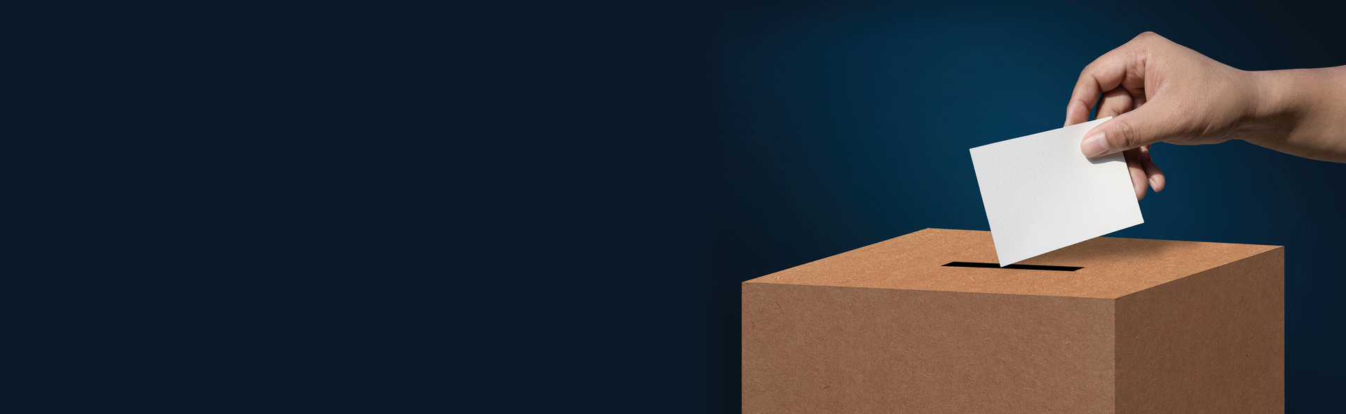 Hand dropping a white ballot into a cardboard voting box against a dark blue background