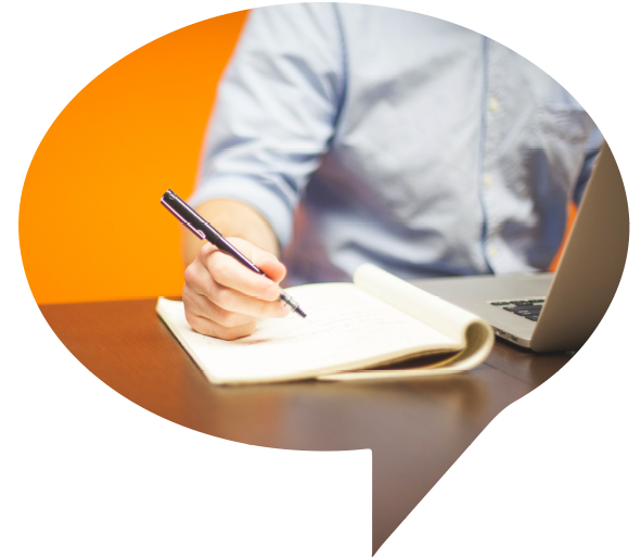 Person writing in a notebook beside a laptop on a desk with a warm orange background
