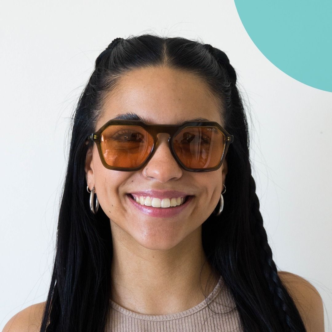 Mujer sonriendo, con gafas de sol con marco marrón y lentes naranjas, parte superior marrón claro y trenzas.