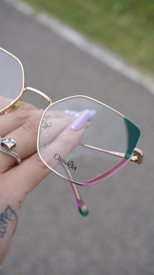 Gafas doradas con detalles en verde y rosa sostenidas en una mano con un anillo de diamantes.