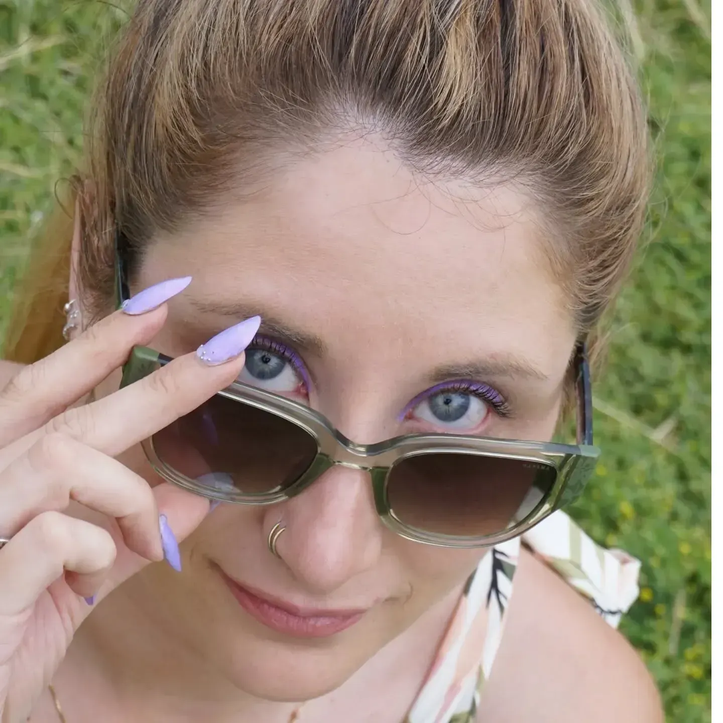 Mujer con ojos azules, uñas color lavanda y gafas de sol, mirando por encima de las gafas.