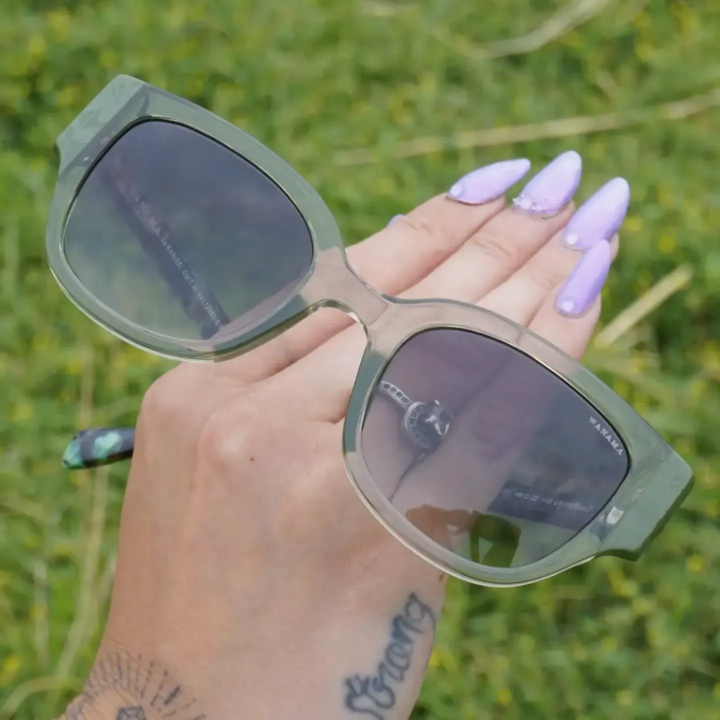 Mano sosteniendo gafas de sol verdes con lentes violetas contra un fondo de césped borroso.