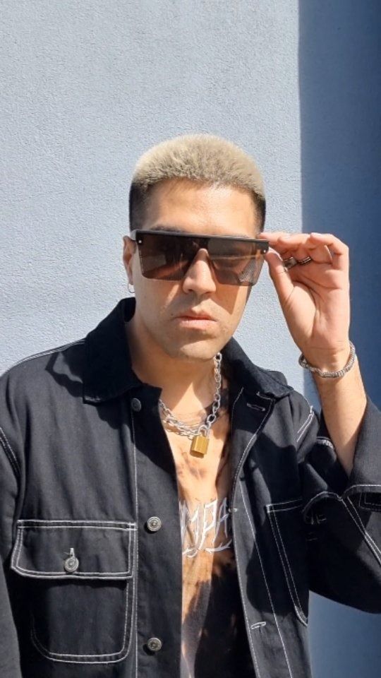 Hombre con chaqueta negra y gafas de sol ajustándose las gafas, de pie frente a una pared azul.