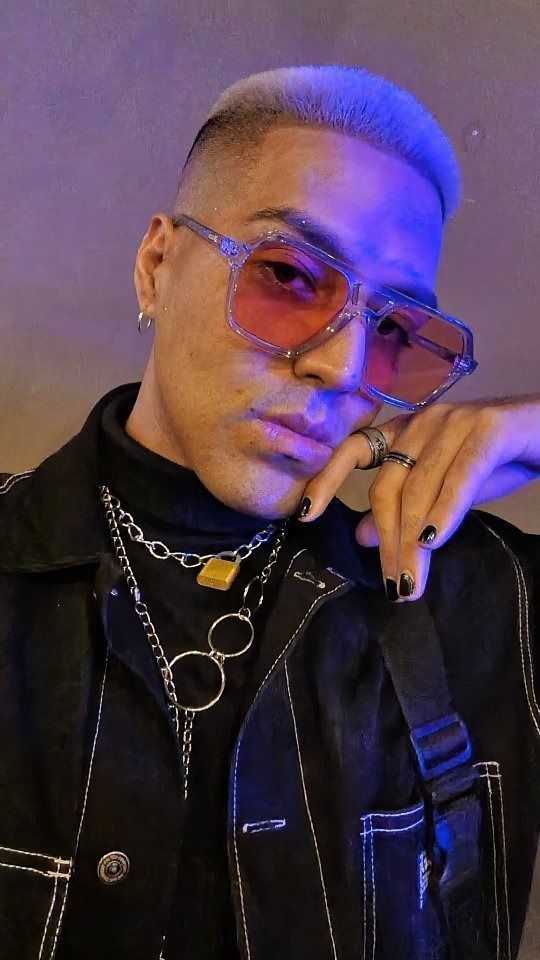 Un hombre con gafas de color rosa, uñas negras y joyas posa frente a una luz azul.