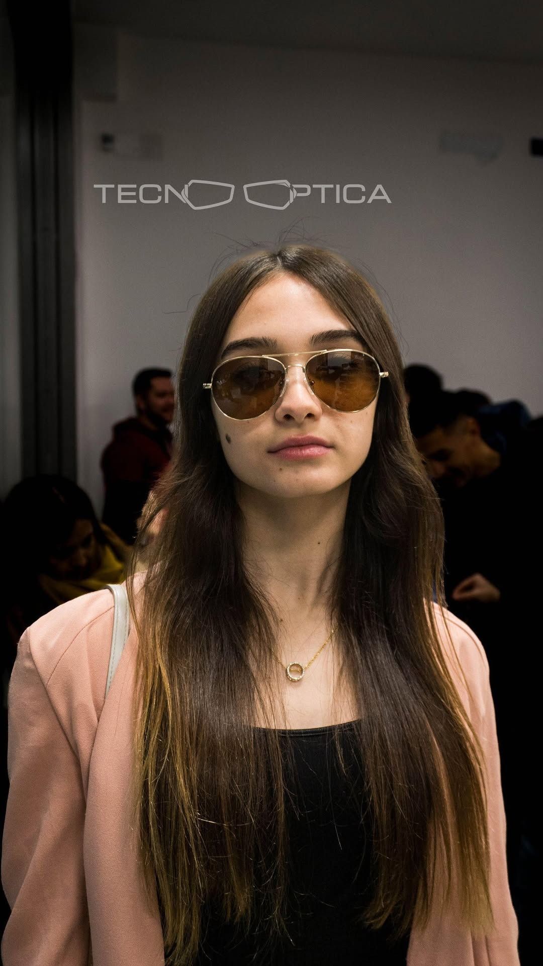 Mujer con gafas de sol, de pie en un interior; fondo borroso con gente. Logotipo de