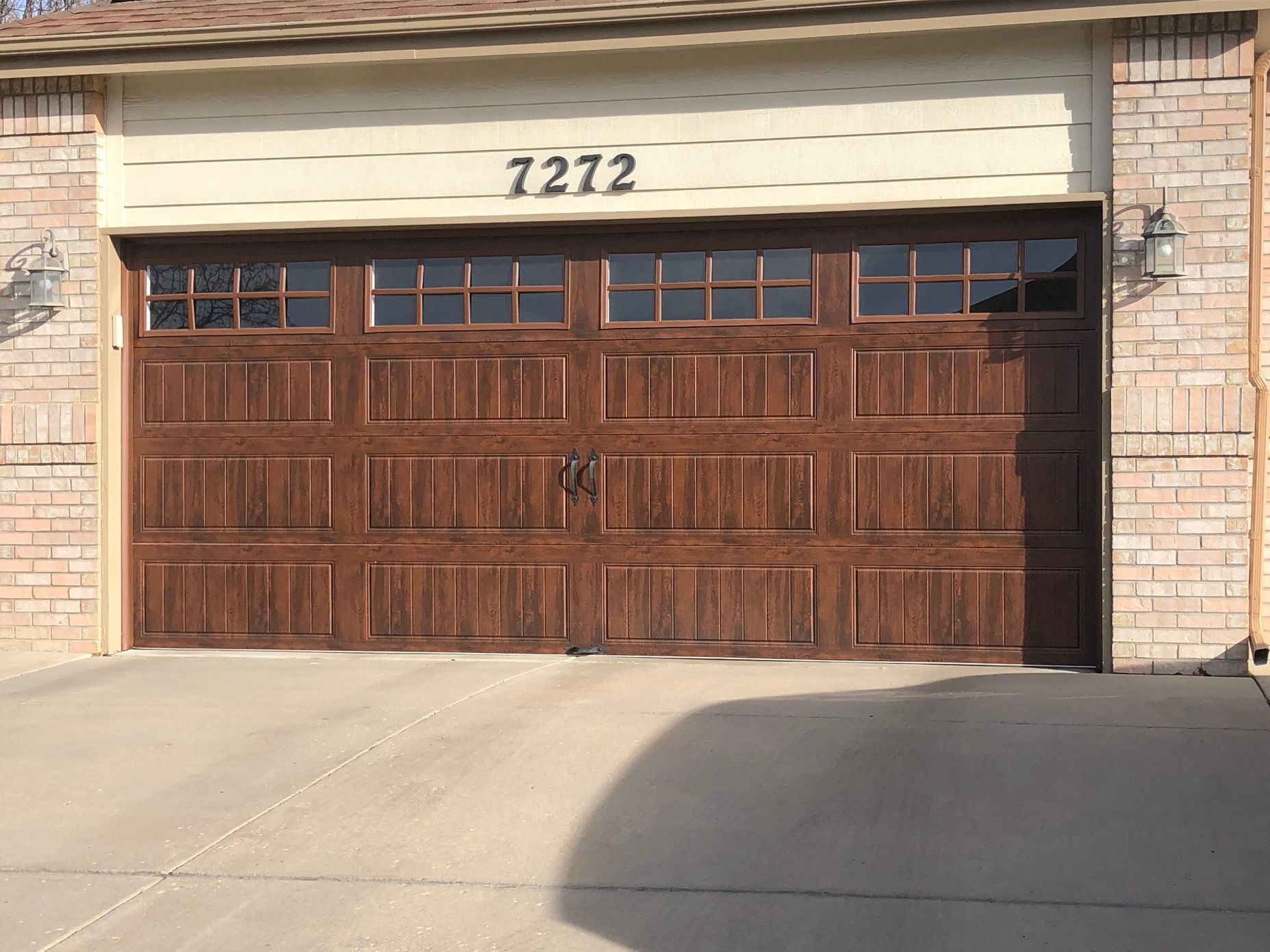 Brown carriage garage door with windows, address 7272 above it, brick exterior, and concrete driveway.