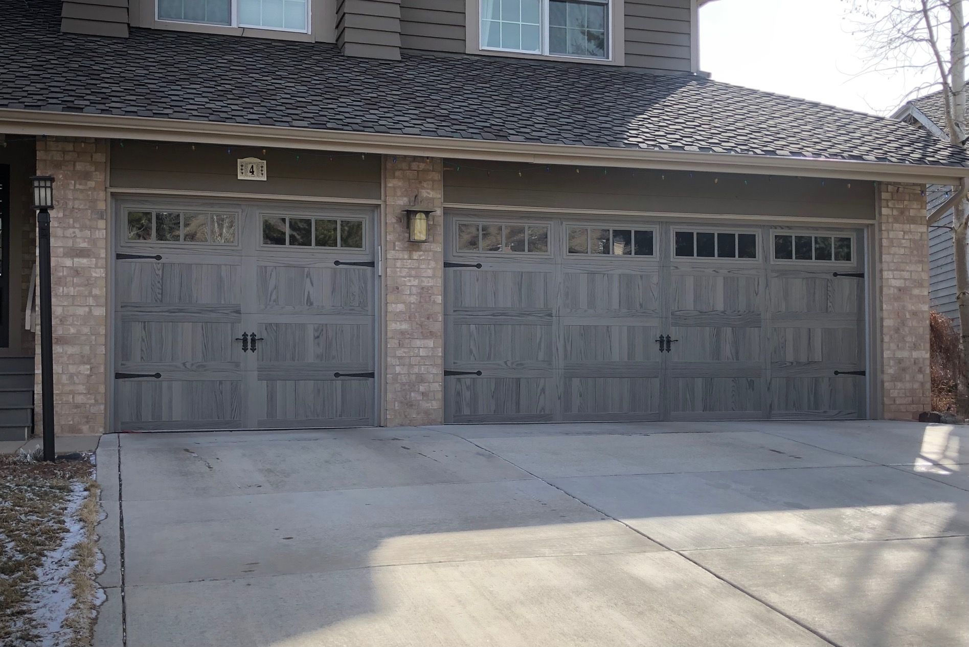 Two gray garage doors with decorative black hardware and brick accents. Concrete driveway.