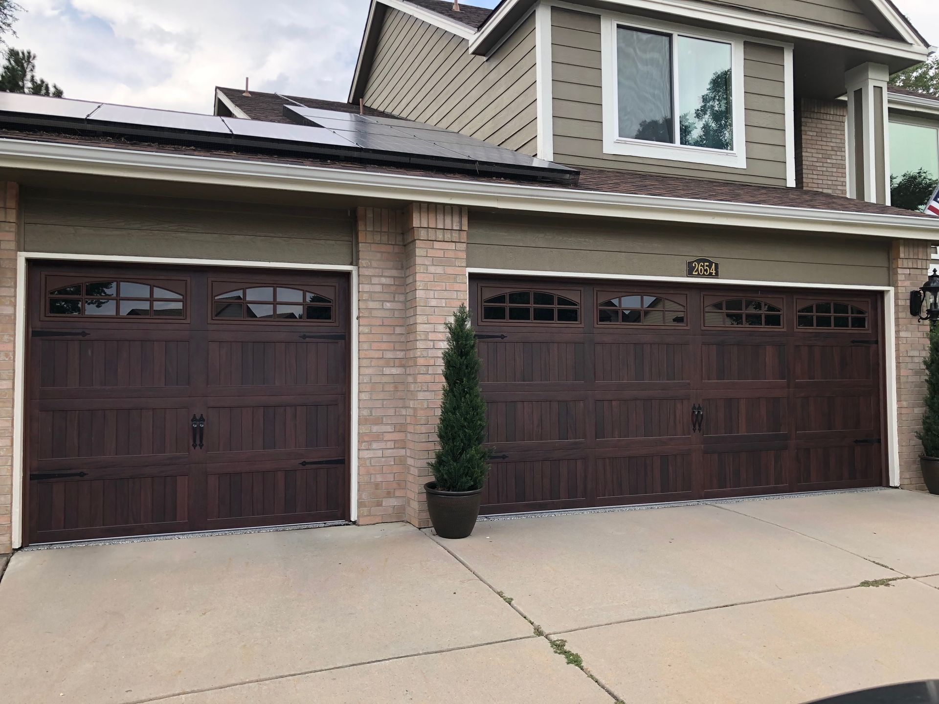 Two-car carriage garage with dark oak doors, tan trim, brick facade, and potted topiary. The house is two-story.