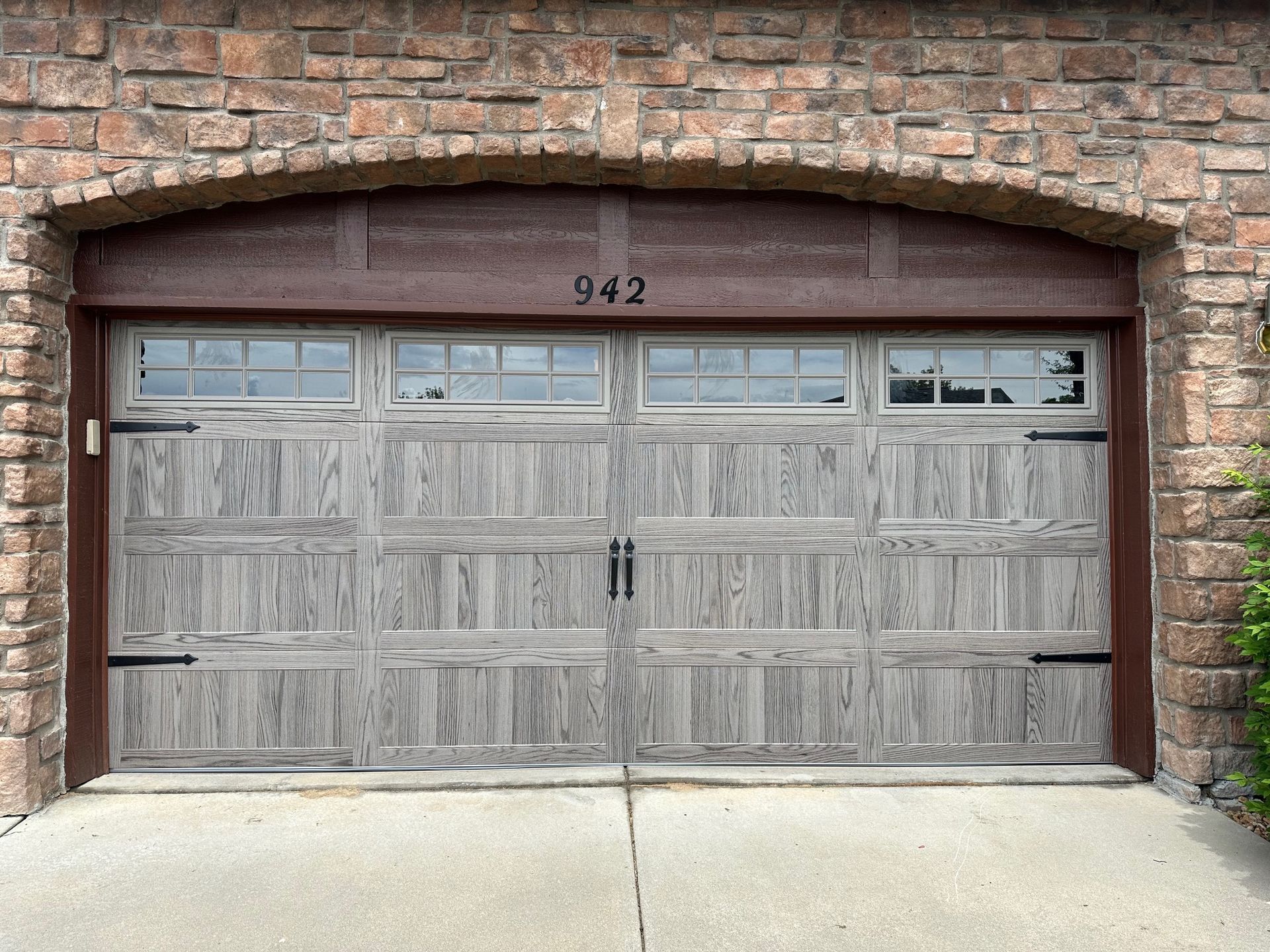 Driftwood carriage garage door with stockton windows, black hardware, and arched brick facade. 