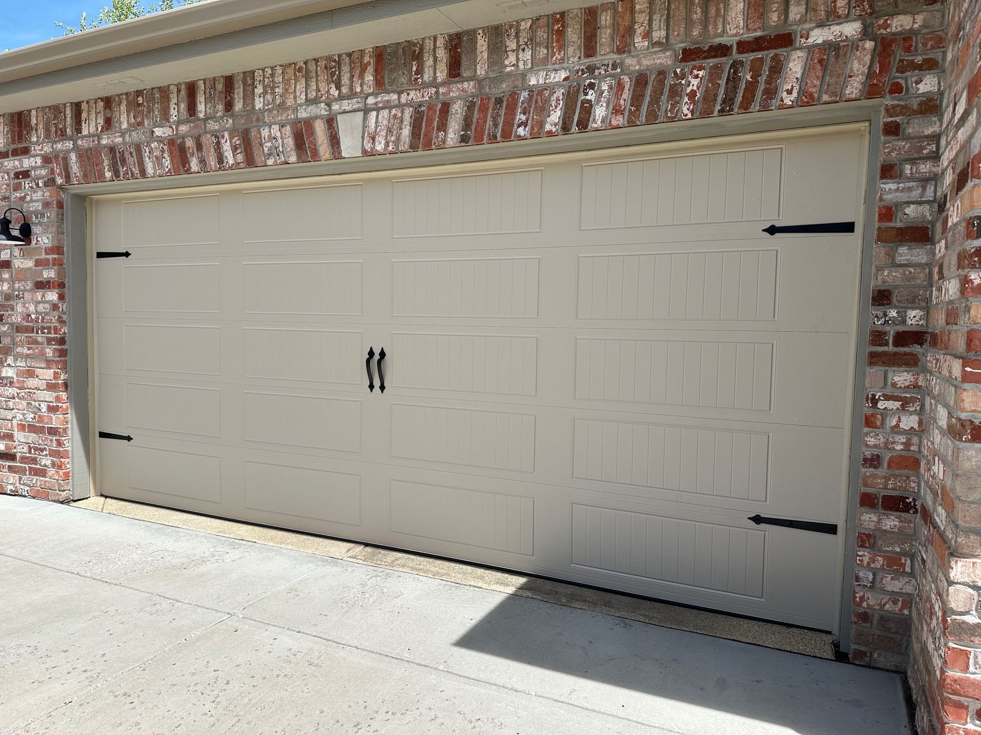 Tan carriage garage door with black hinges and handles, set in a brick building.