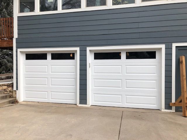 Two white garage doors with windows under a blue-gray house, concrete driveway.