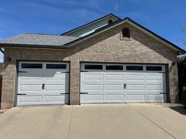 Two white garage doors on a brick house, black hardware, concrete driveway, blue sky.