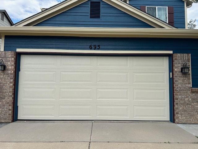 Beige garage door on a blue house with address number 695. Brick columns flank the door.