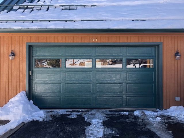Green garage door with windows; snow on the ground and roof. Wooden exterior.
