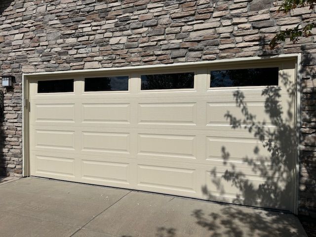 Beige garage door with windows, set against a stone wall and concrete driveway.