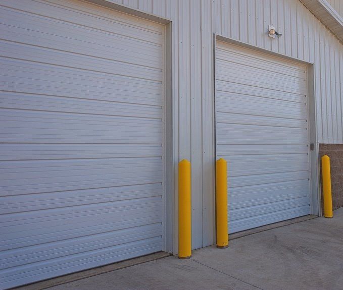 Two white garage commercial doors with yellow bollards outside a building.