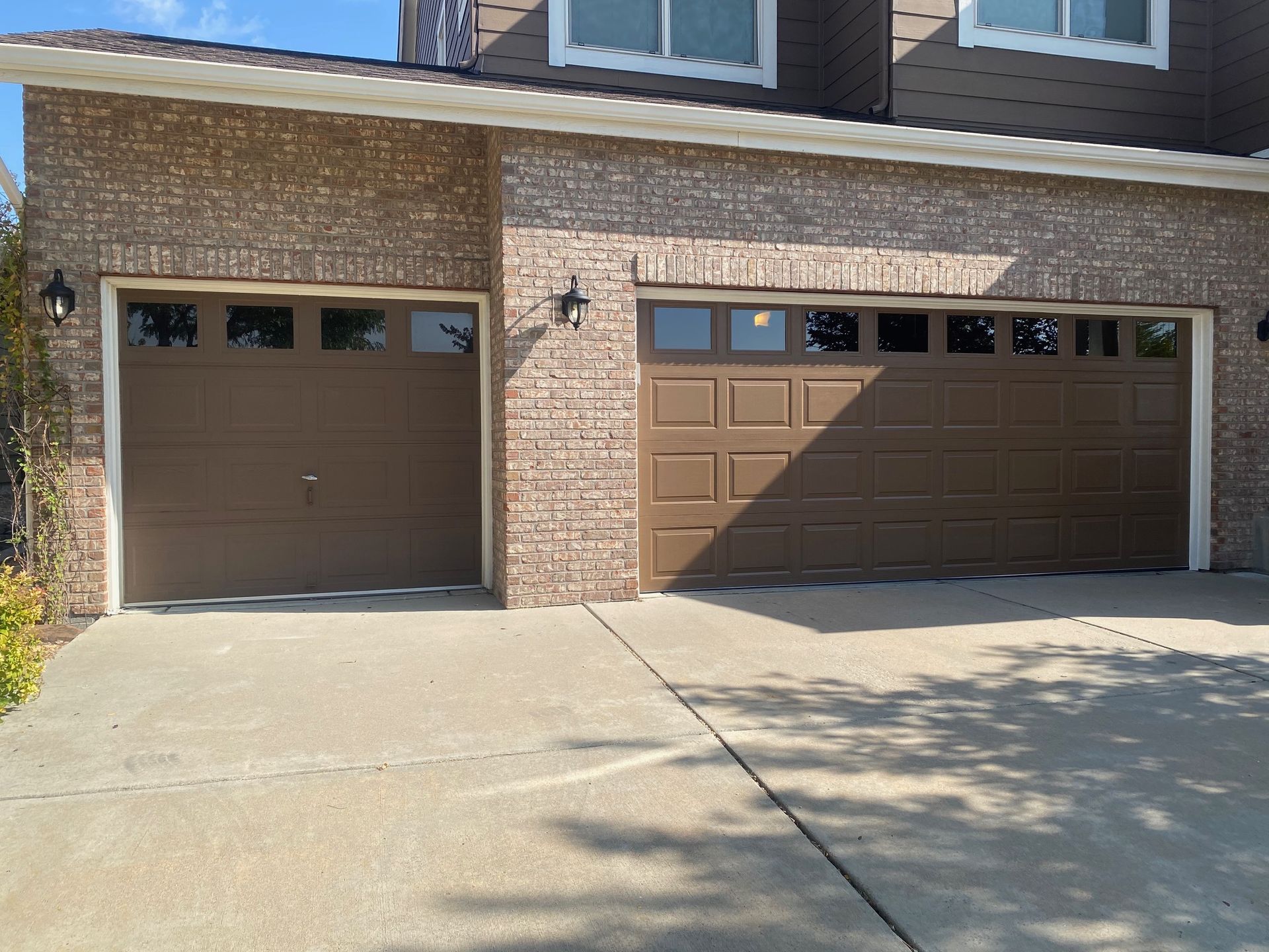 Two brown garage doors in a brick building. Sunlight casts shadows on the concrete driveway.