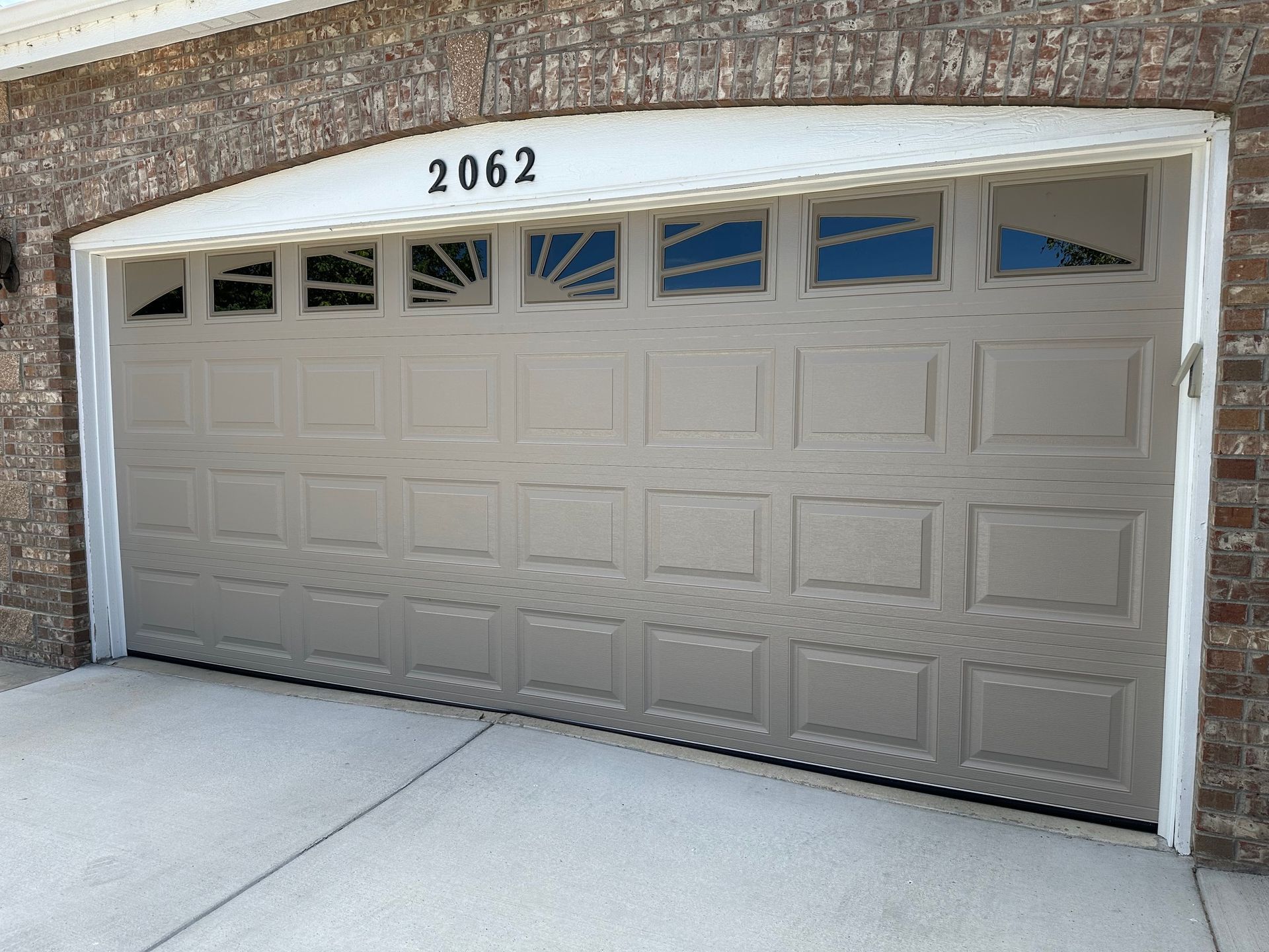 Tan garage door with rectangular panels, address 2062 above, brick and concrete setting.
