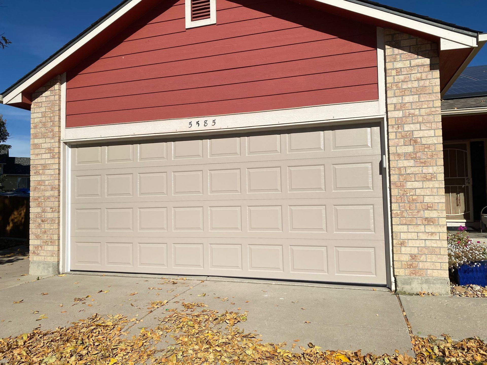 Tan garage door with red siding and brick accents. Fall leaves on the ground.