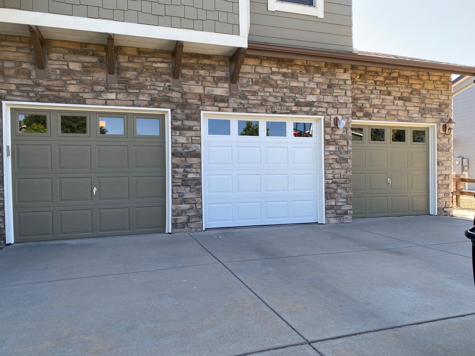 Three garage doors, two gray and one white, on a stone building, concrete driveway.