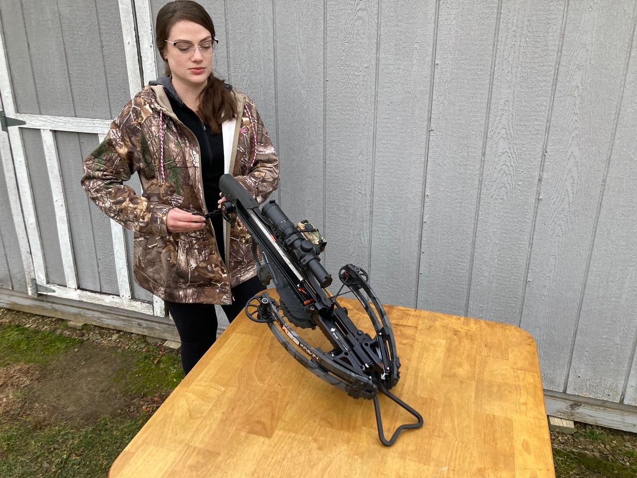 Woman in camouflage jacket with a crossbow on a wooden table.