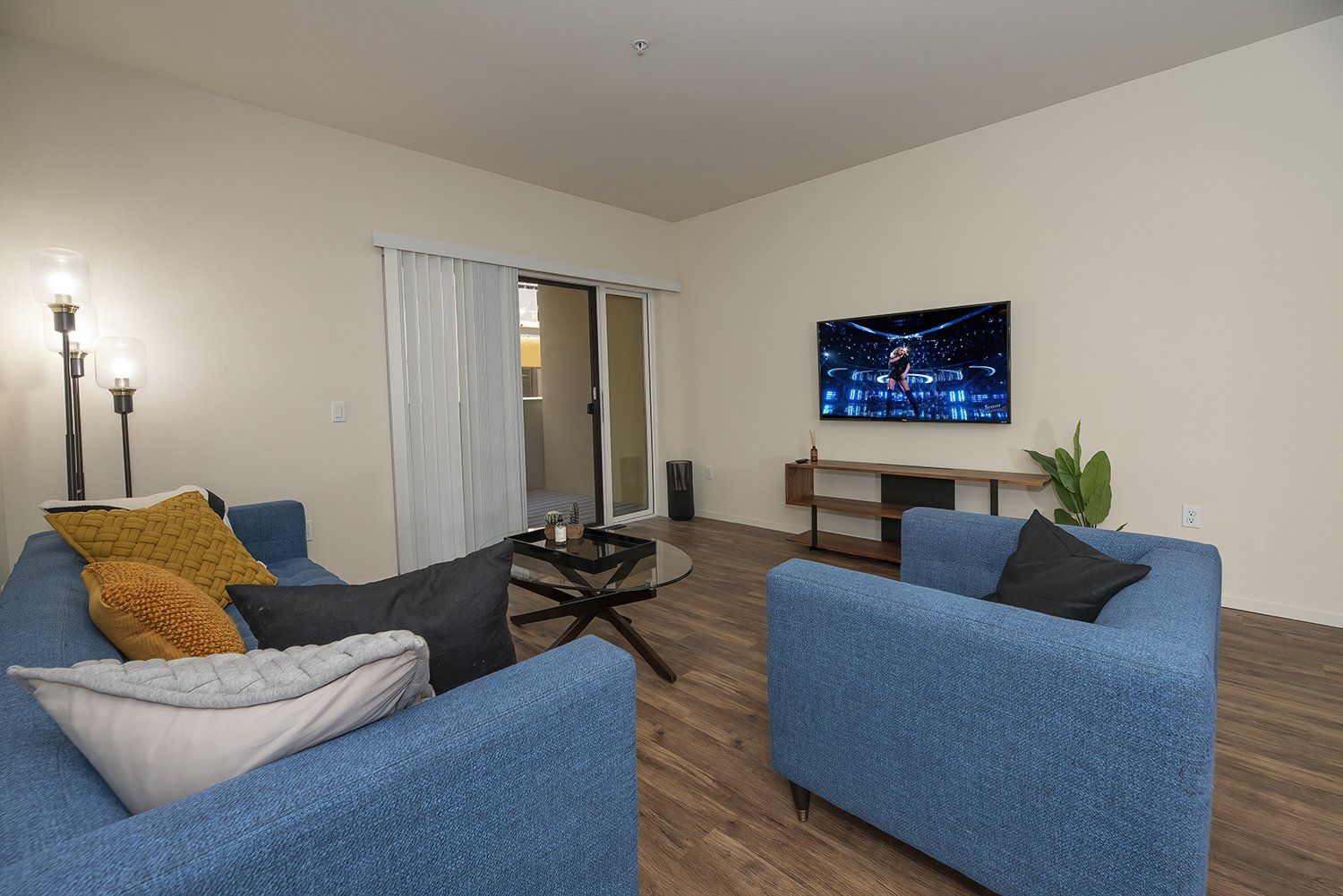 Apartments for Rent in El Cerrito CA - Metro 510 - Living Room with Wood-Style Flooring, Blue Sofa and Armchair, Mounted TV, Storage Shelving, Beige Walls, Standing Lamps, Glass-Topped Coffee Table, and Sliding Glass Door