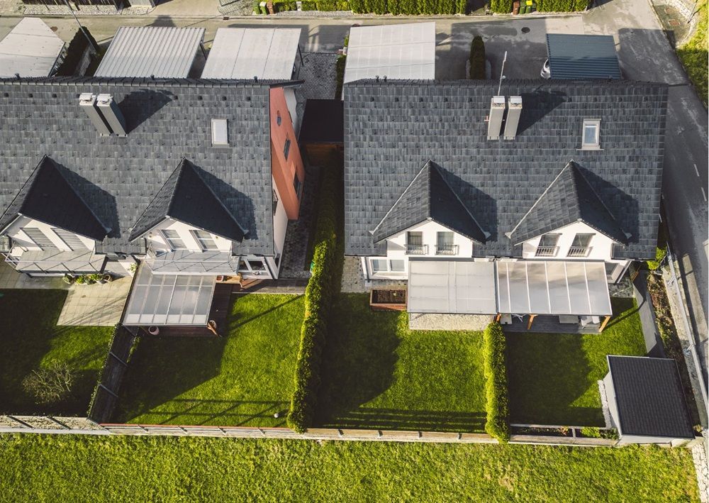 Overhead drone view of modern semi-detached homes with dark roofs, white garages, solar panels, and green backyards.