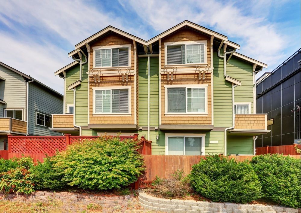 Sage green craftsman style duplex with wood shingle accents, peaked roofs, white windows, and landscaped front yard with shrubs.