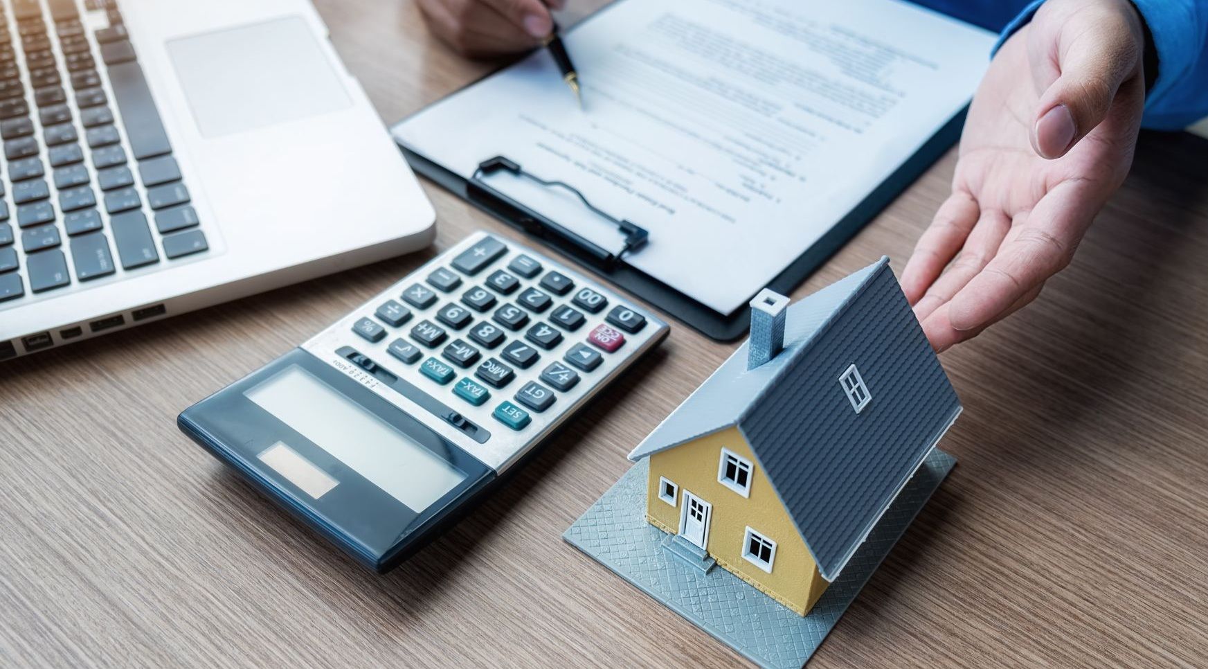Close-up of a desk with a laptop, calculator, clipboard holding a document, and a small yellow house