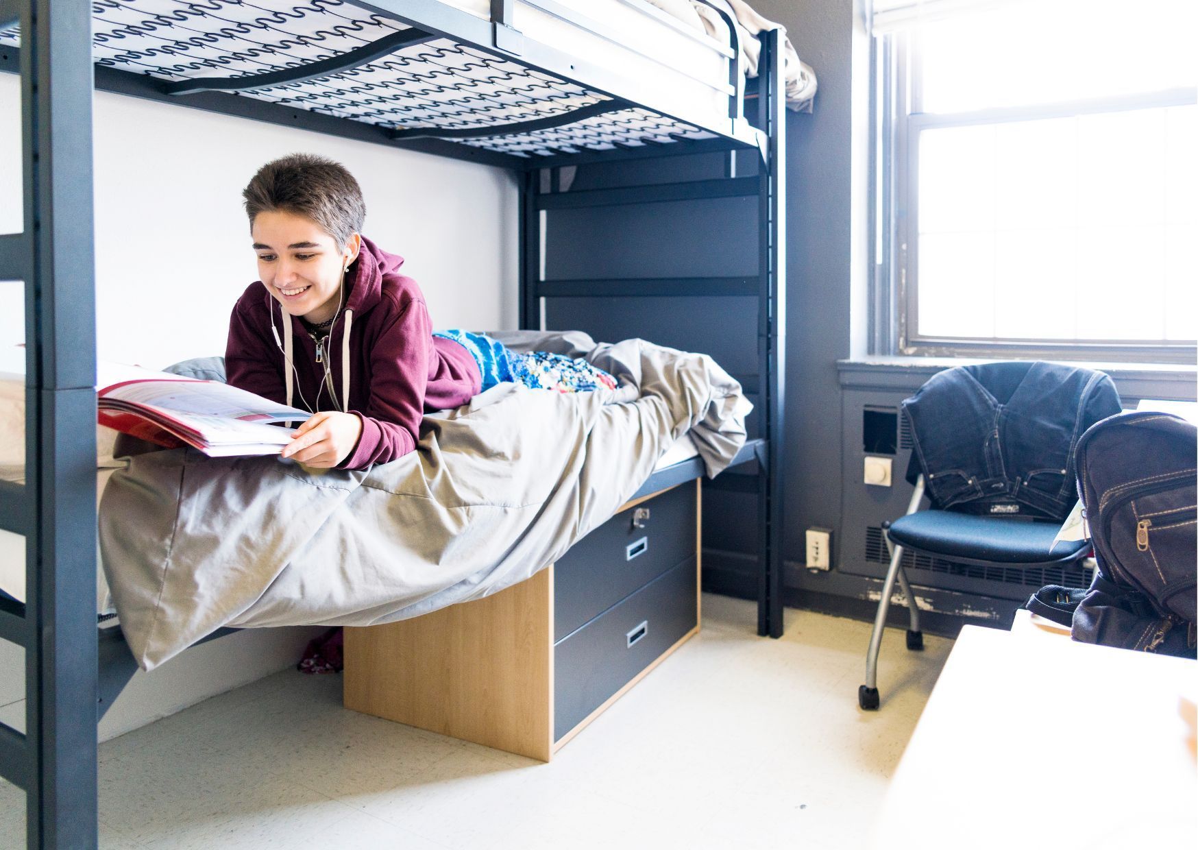  Student in maroon hoodie smiling while reading book on lower bunk bed in dormitory room.