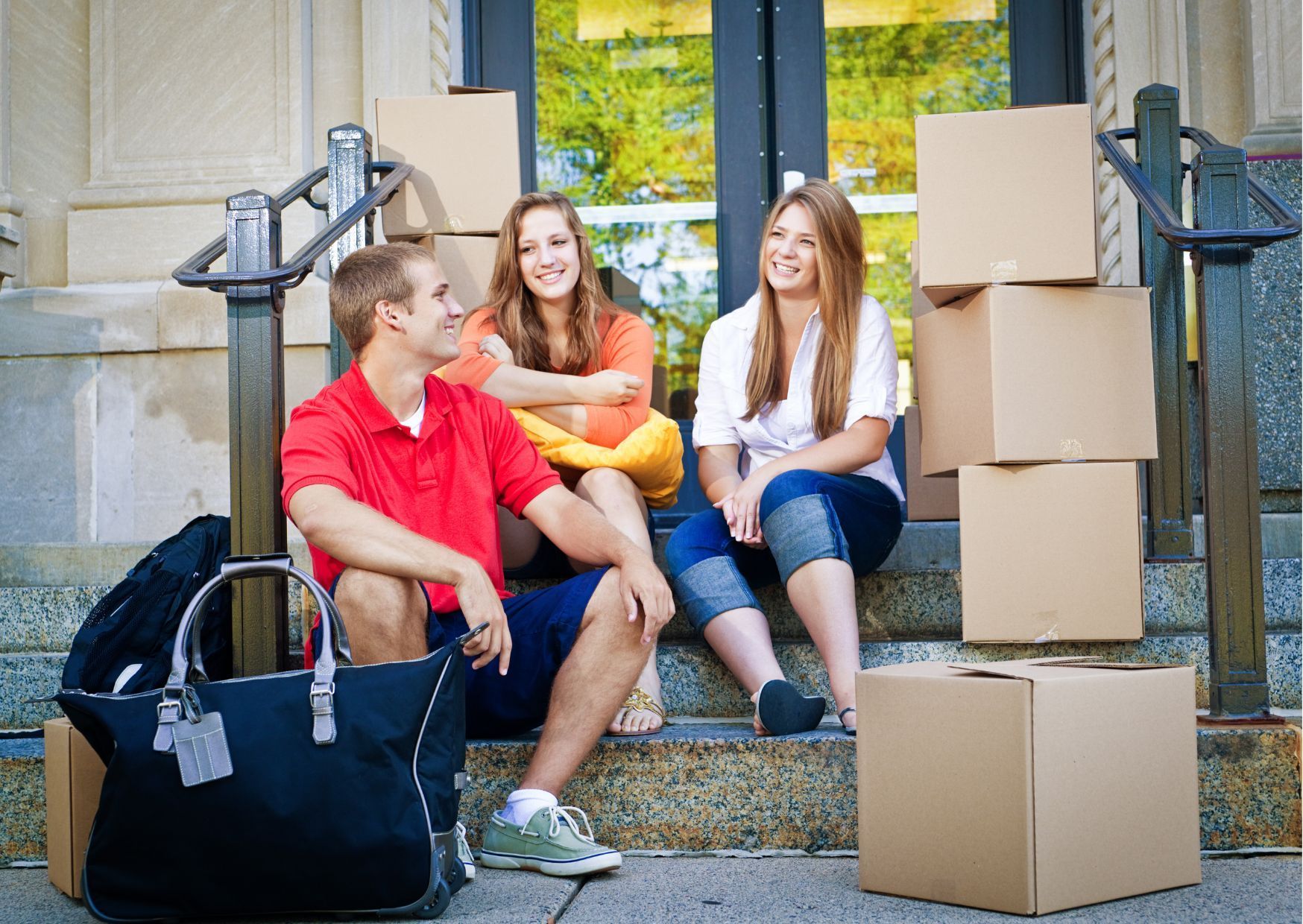 Three individuals sitting on stone steps surrounded by cardboard boxes and duffel bag outside dormitory entrance.