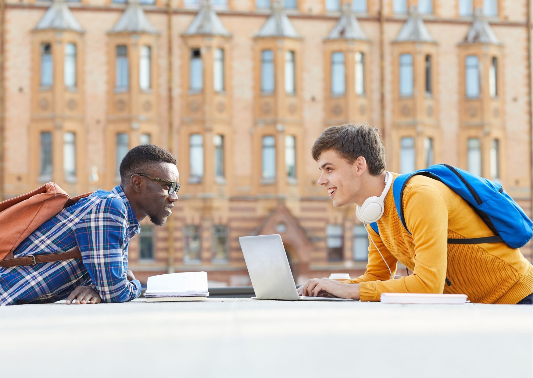Two students with backpacks and laptop studying at outdoor table in front of ornate brick university building.