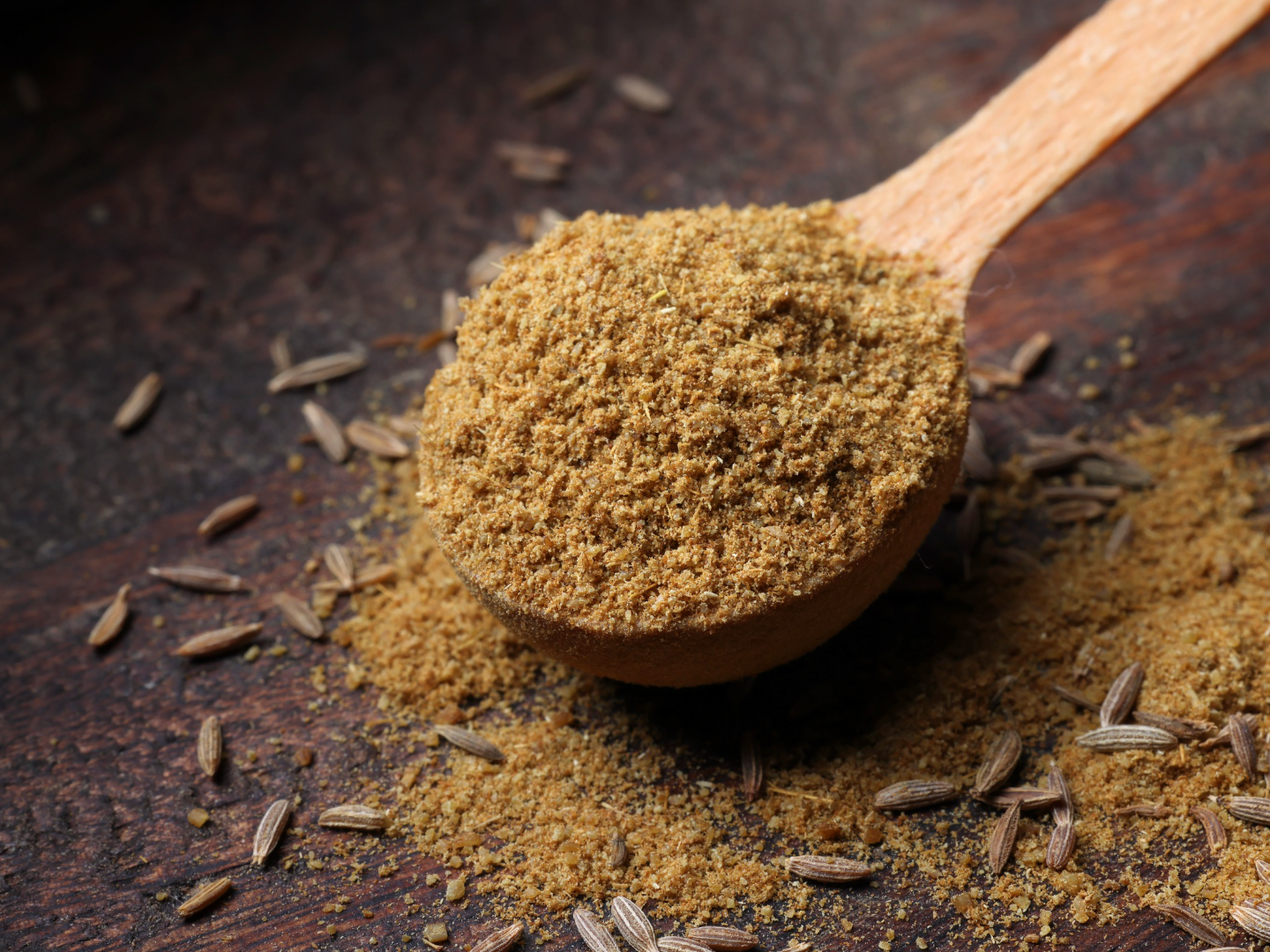 Wooden spoon holding ground cumin, with whole cumin seeds scattered on a wooden surface.