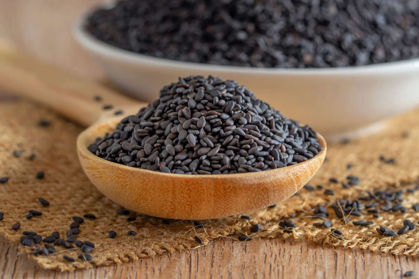 Black sesame seeds in a wooden spoon and a bowl, on burlap and a wooden surface.