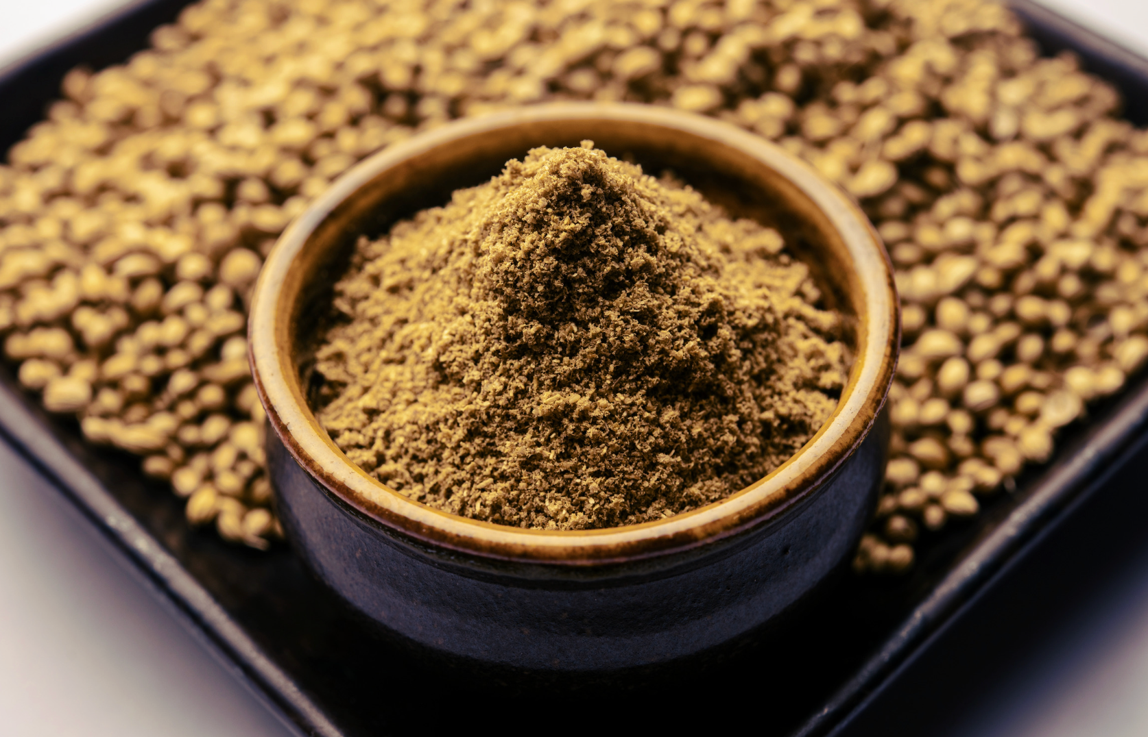 Bowl of ground coriander powder, surrounded by whole coriander seeds on a black tray.