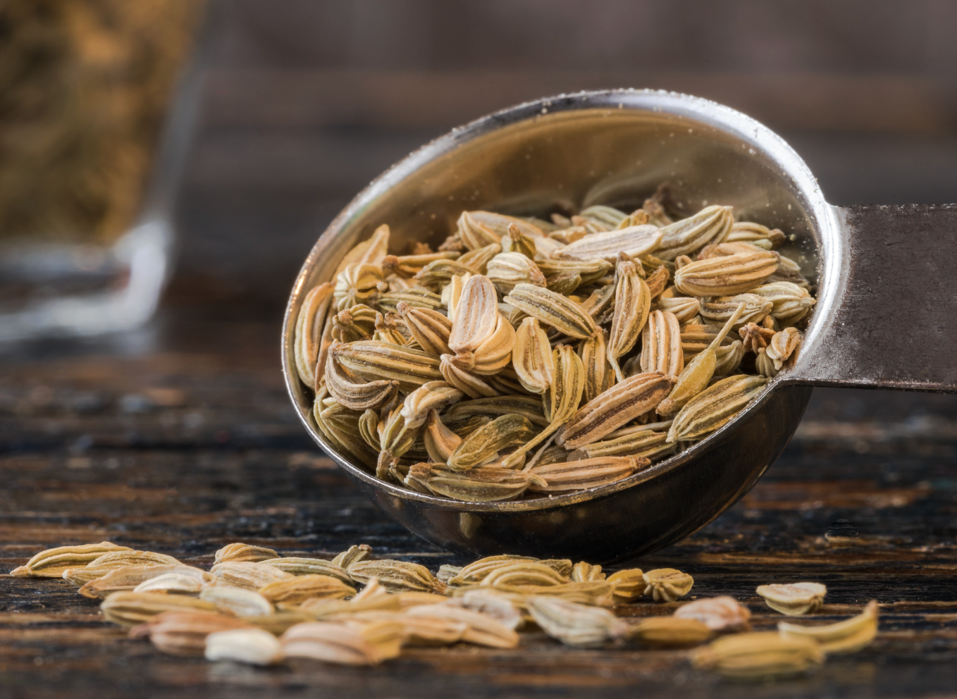 Teaspoon of fennel seeds spilling onto a wooden surface.