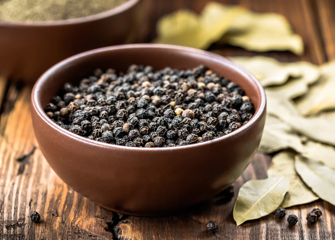 Brown bowl filled with black peppercorns on a wooden surface, with bay leaves visible.