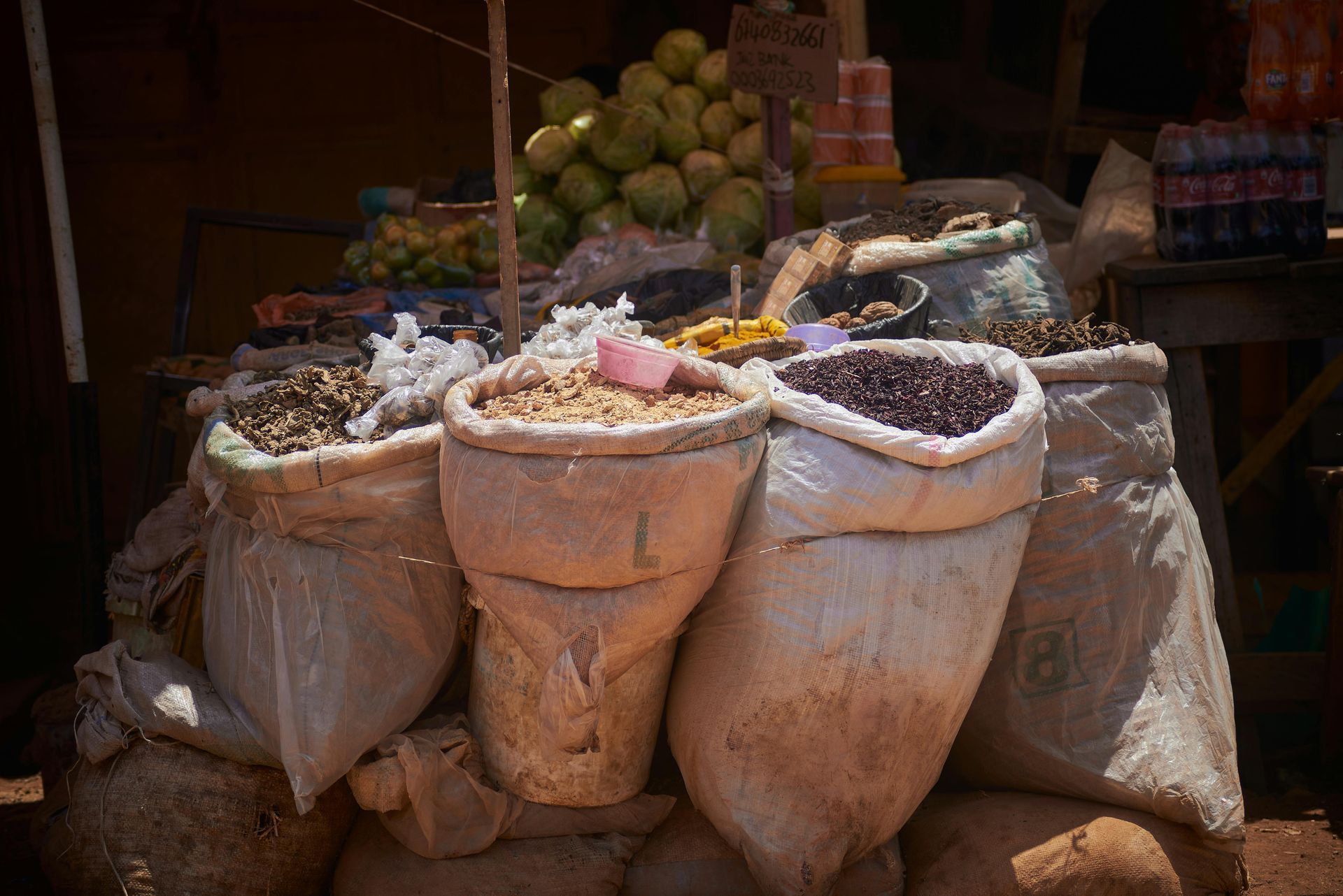 Bags of grains and seeds at an outdoor market stall, with fruit and other goods in the background.