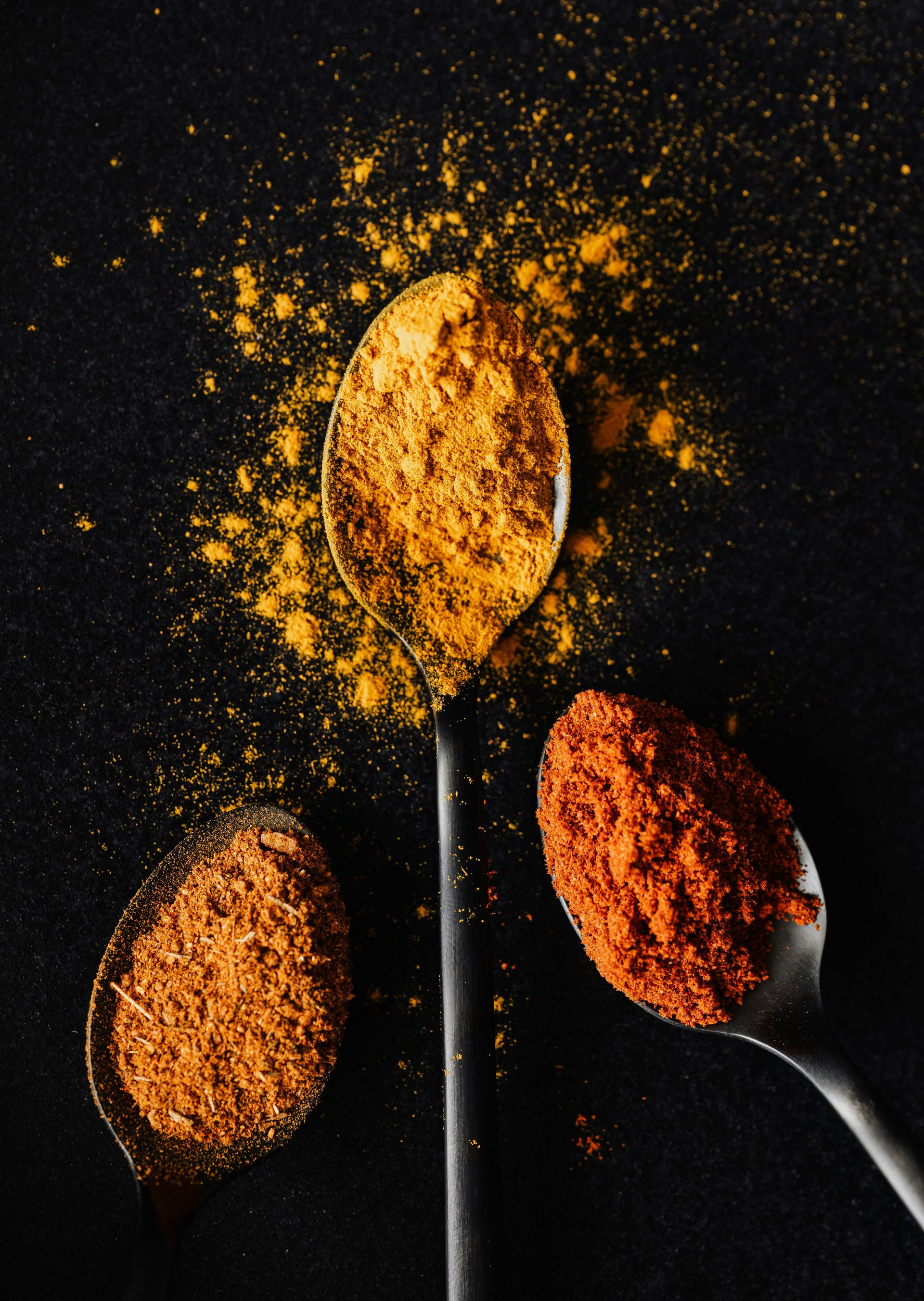 Three spoons holding orange and red spices, scattered on a black surface.