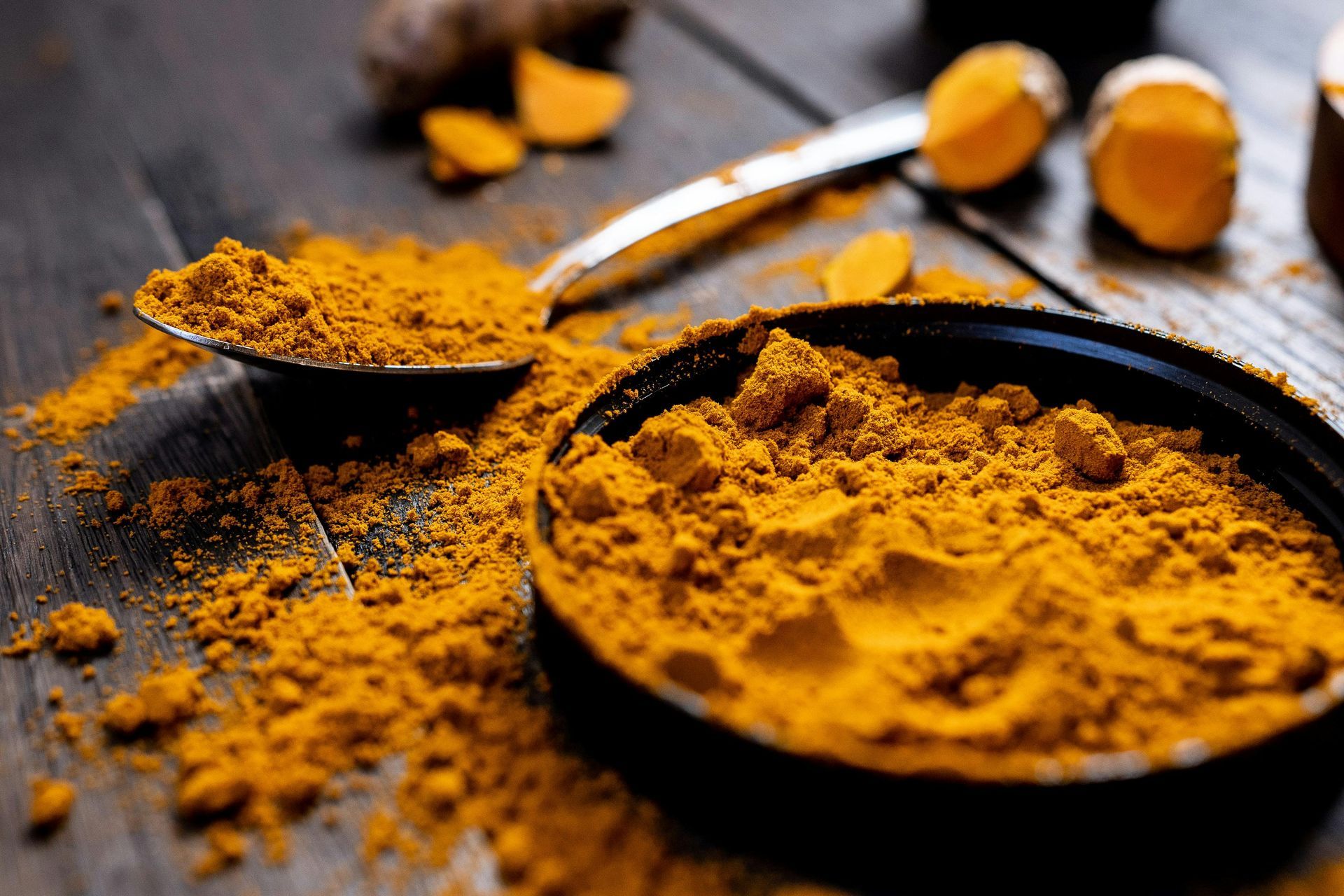 Close-up of turmeric powder in a black container with a spoon and scattered turmeric pieces on a wooden surface.