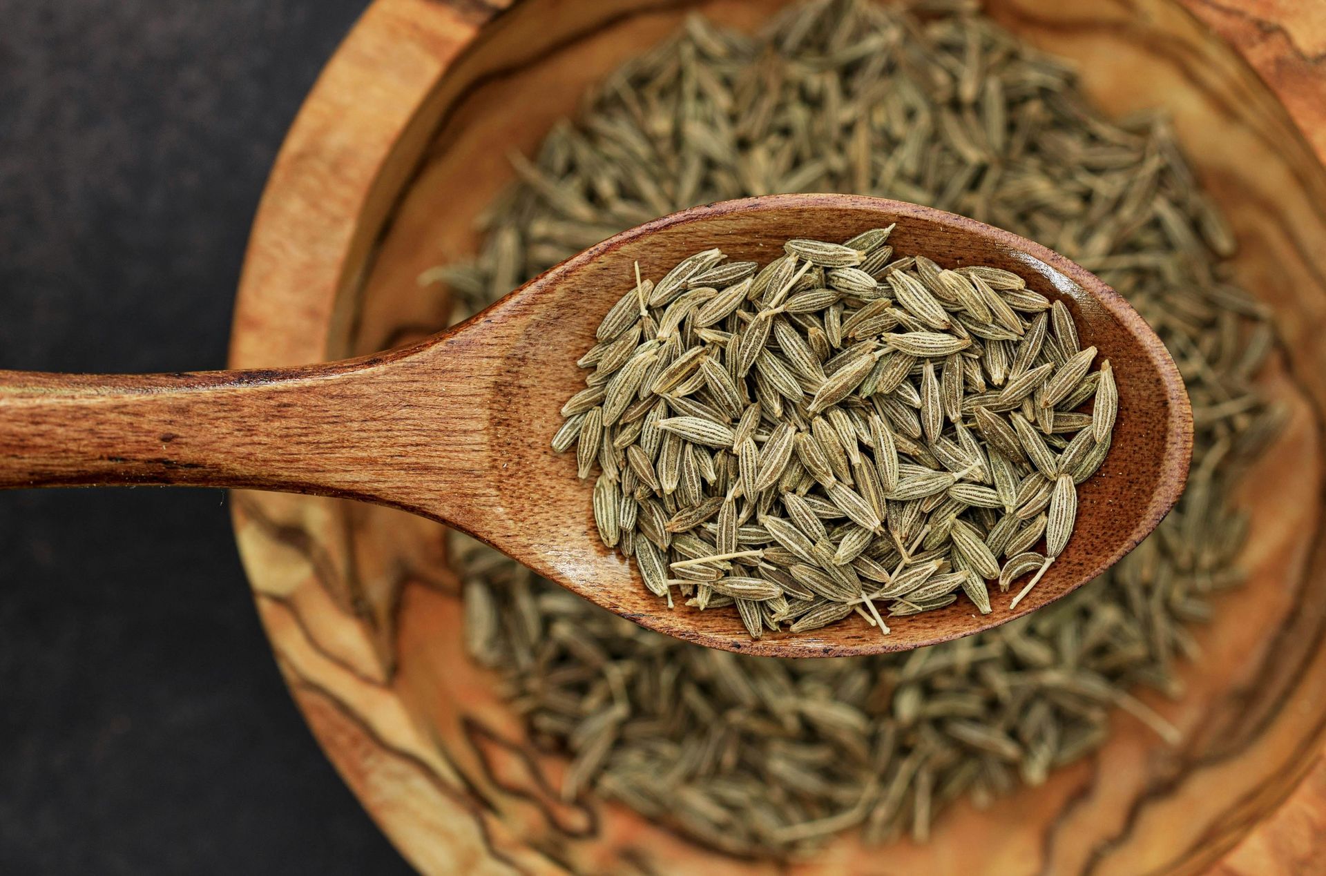 Wooden spoon holding cumin seeds, over a wooden bowl filled with seeds.