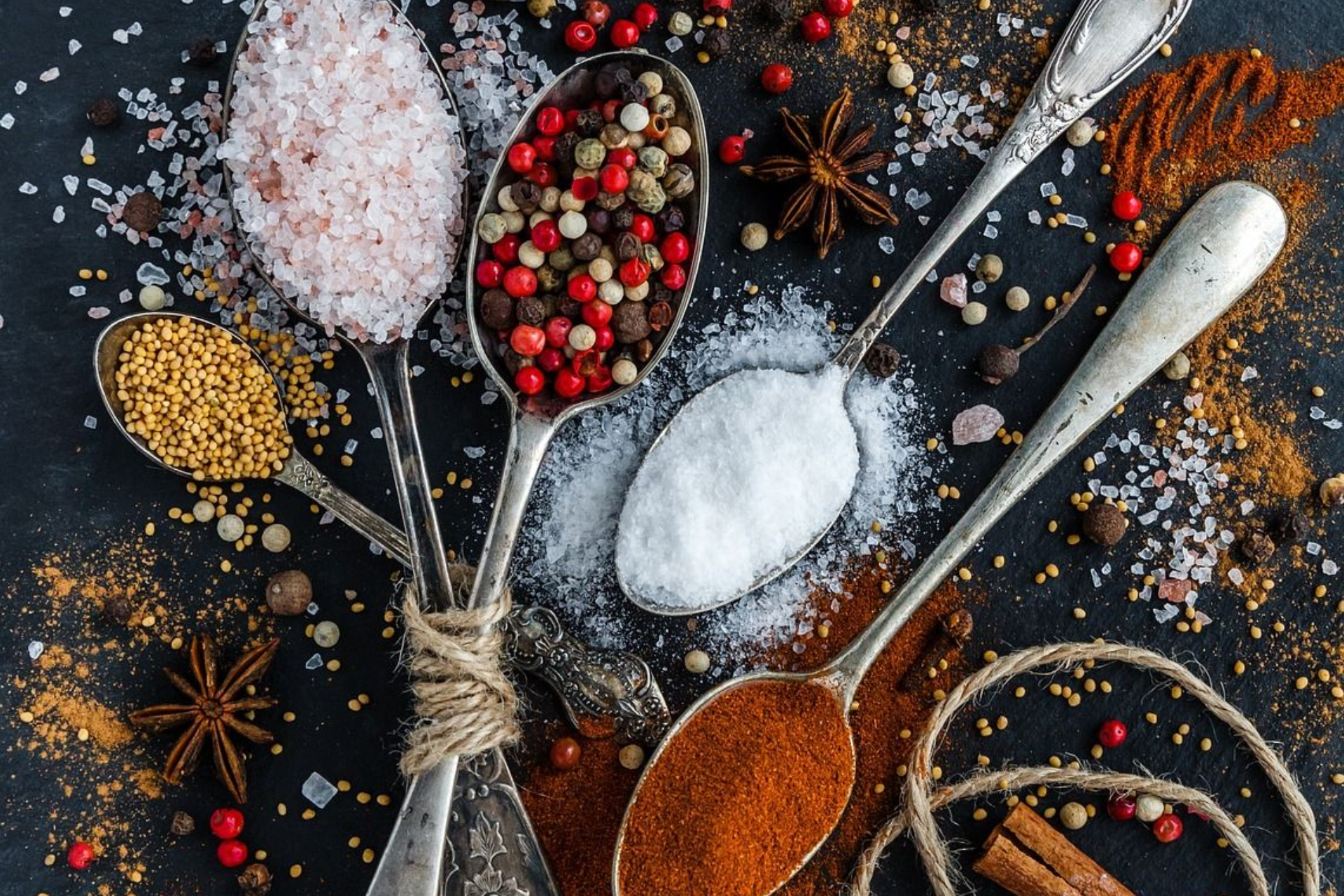 Teaspoons filled with various spices, including salt, pepper, and chili powder, arranged on a dark surface.