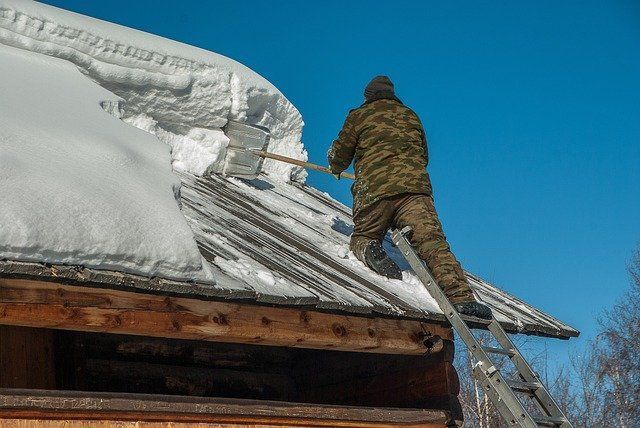 A person in camouflage clothing shovels snow from a rooftop, using a ladder on a clear, sunny day.