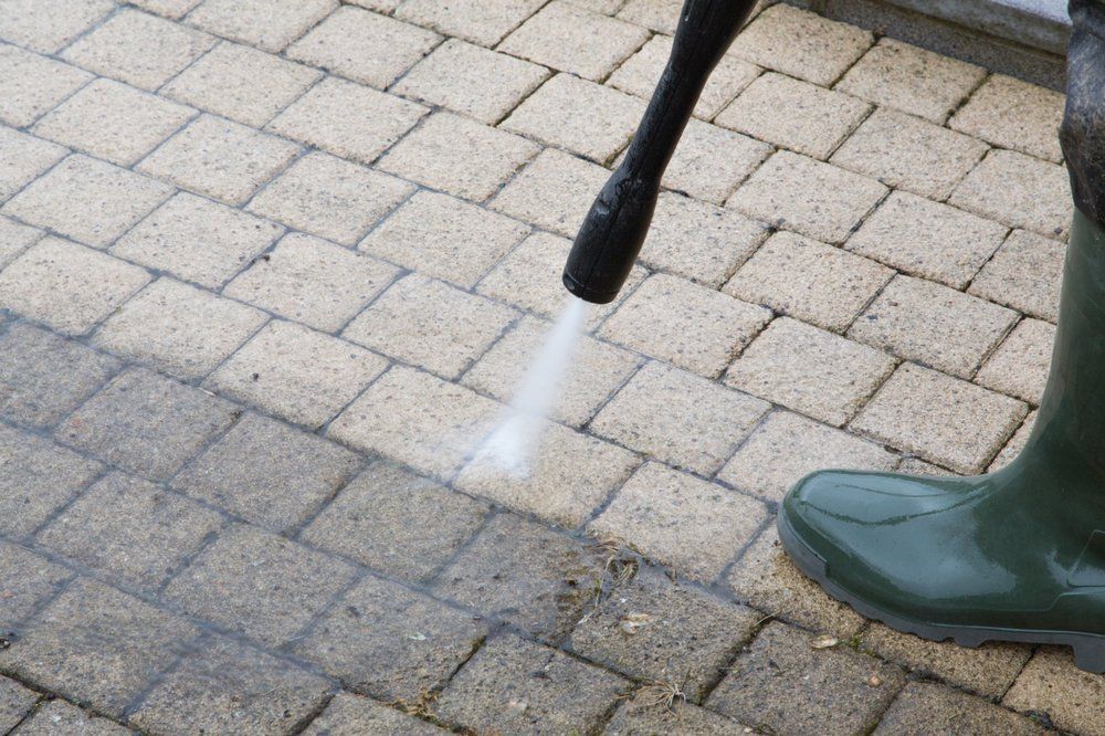 Person in green boots power washing a brick patio, cleaning it from dirty to clean.