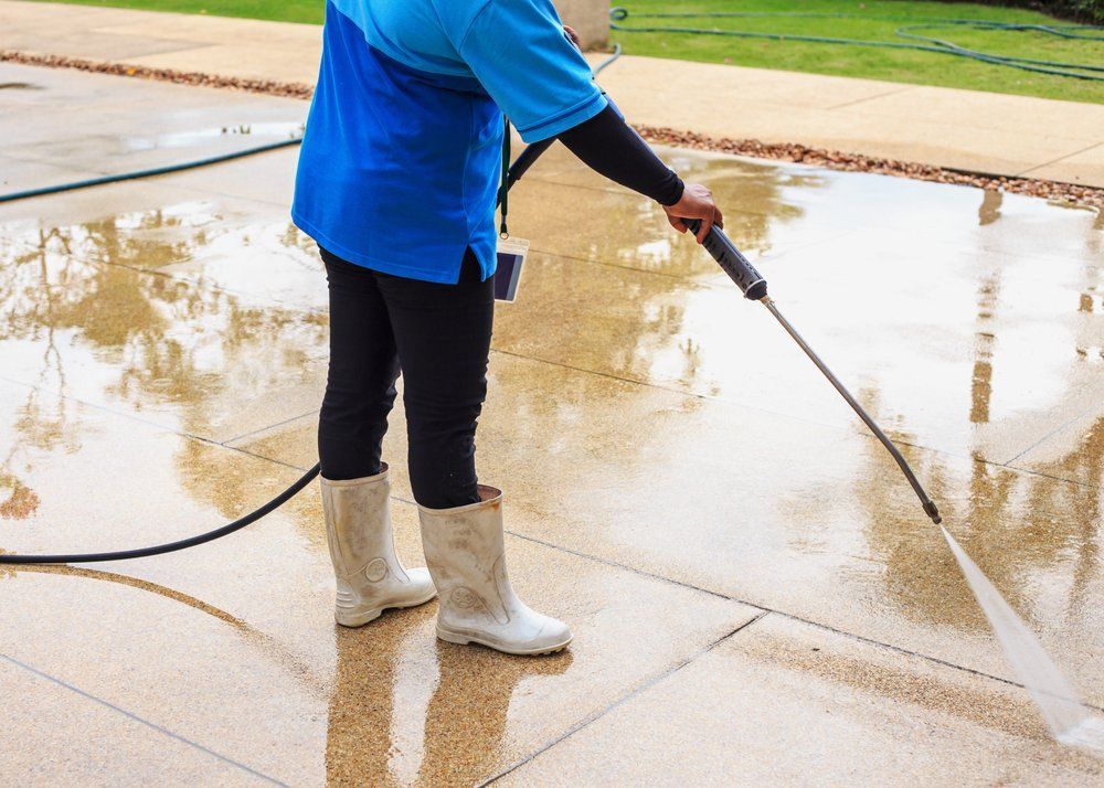 Person in blue shirt and black pants power washes a wet stone patio, wearing white boots.