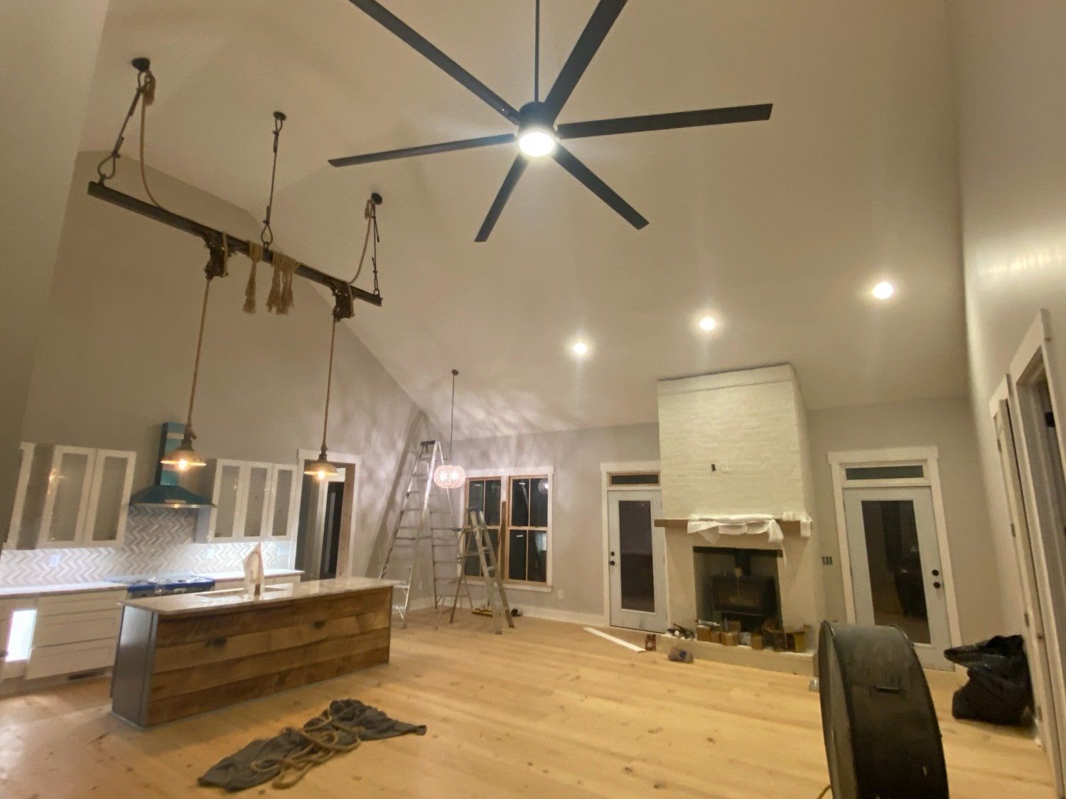 Open-plan kitchen and living area with light wood floors, white walls, a fireplace, and a large ceiling fan.